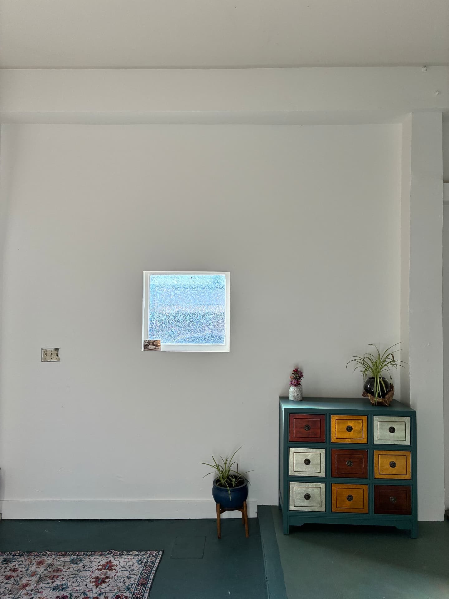 A bright plaster wall with a single framed photograph above a many-coloured painted apothecary chest