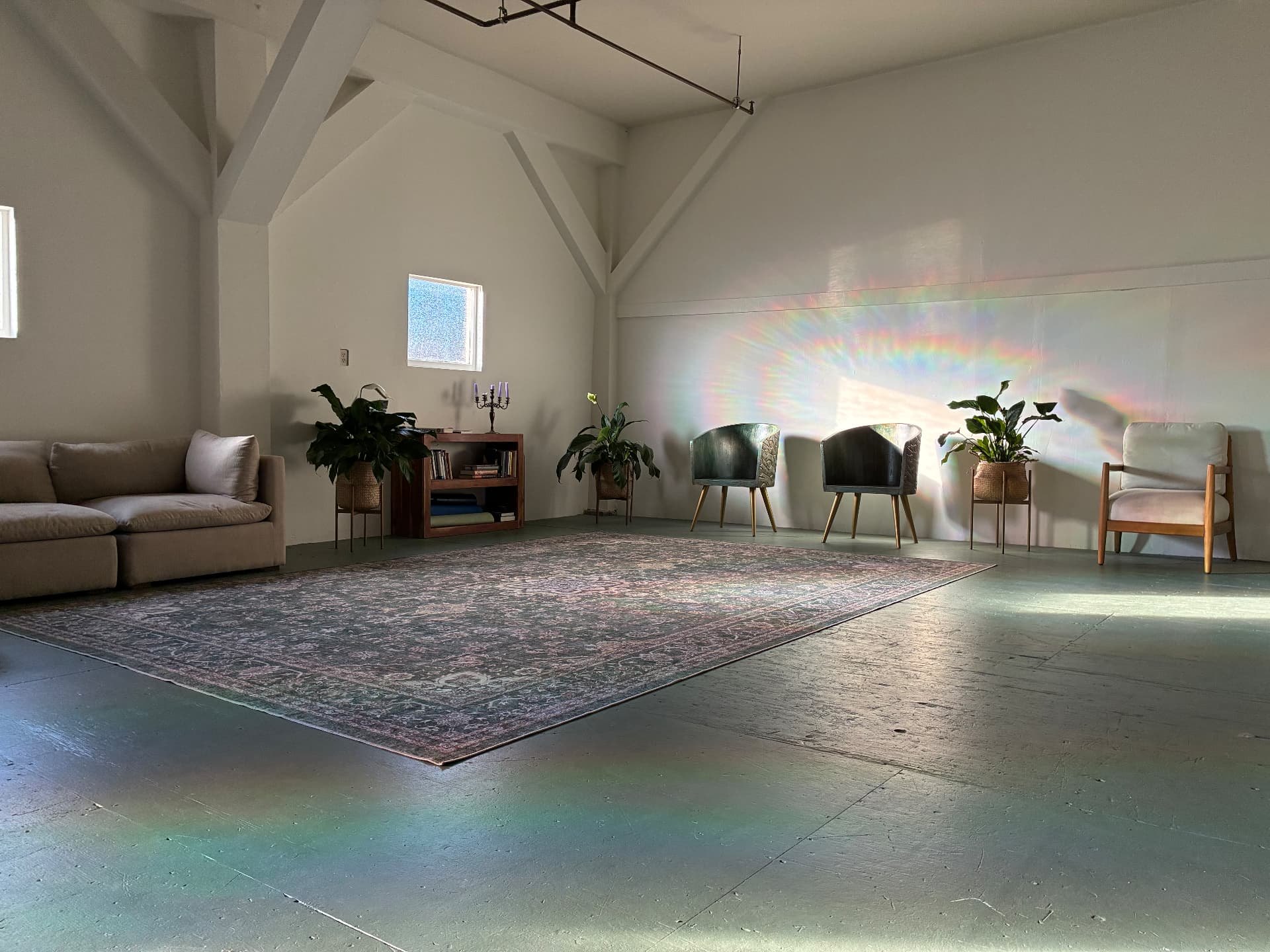 The Harbor empty and waiting: a single rug and armchairs on the green painted floor, rainbow window light across the plaster wall