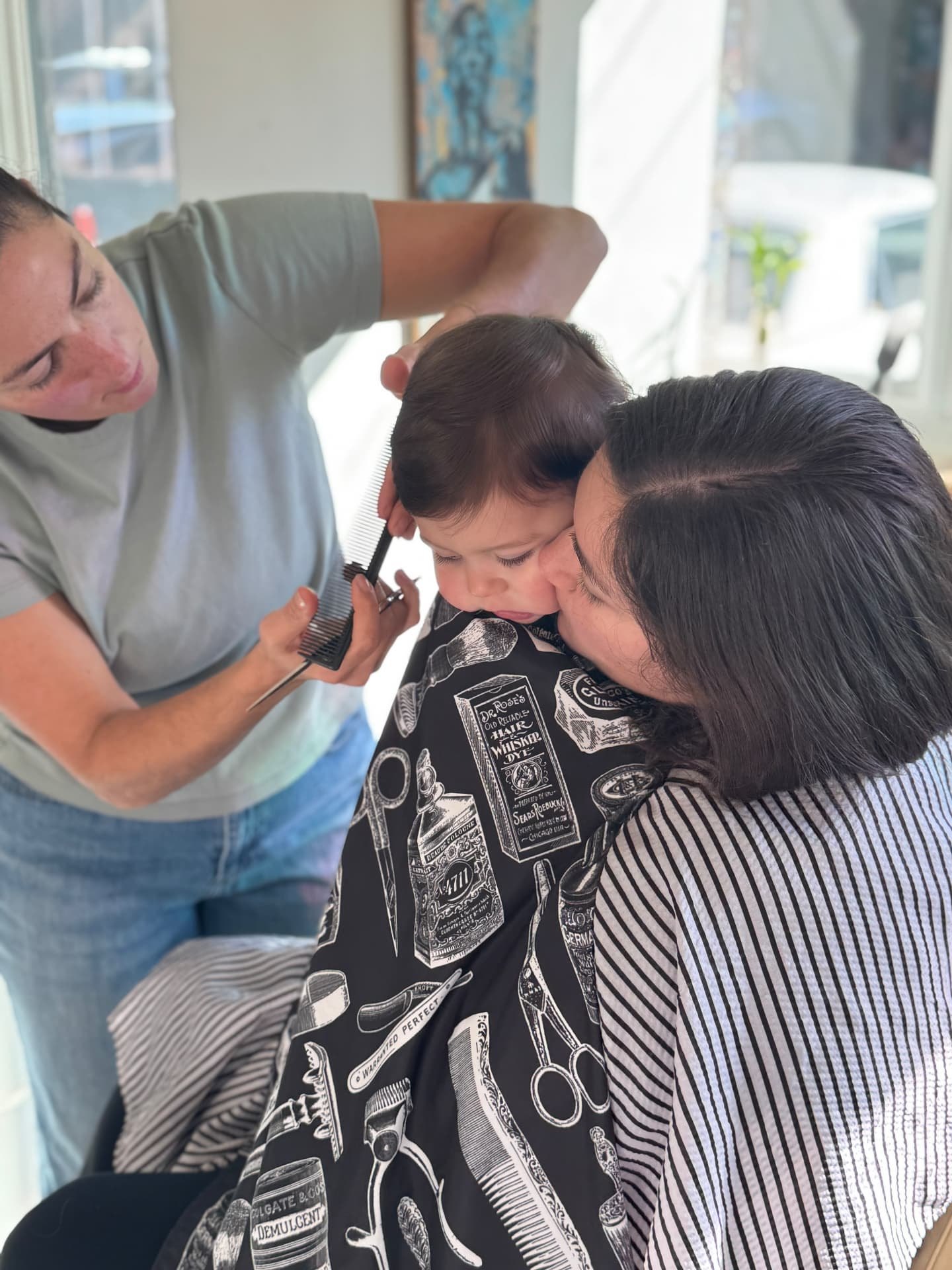 Natasha combing a baby's hair for a first haircut while the baby is held in a parent's lap under a barber cape
