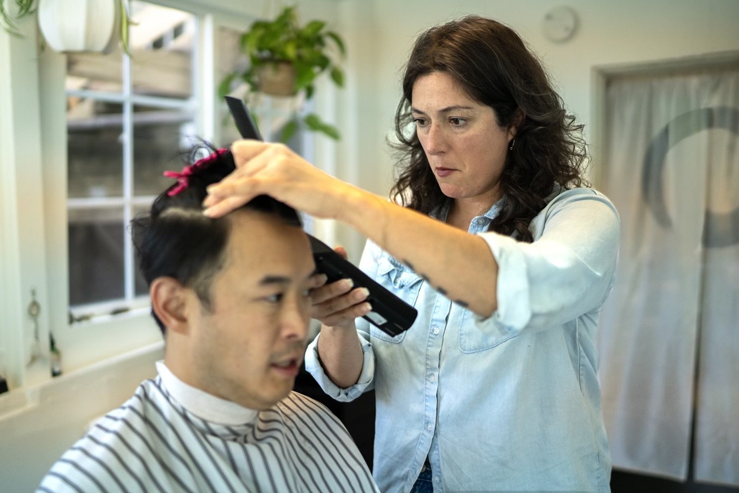 Natasha holding a comb and clippers, concentrating on a client's fade in bright window light