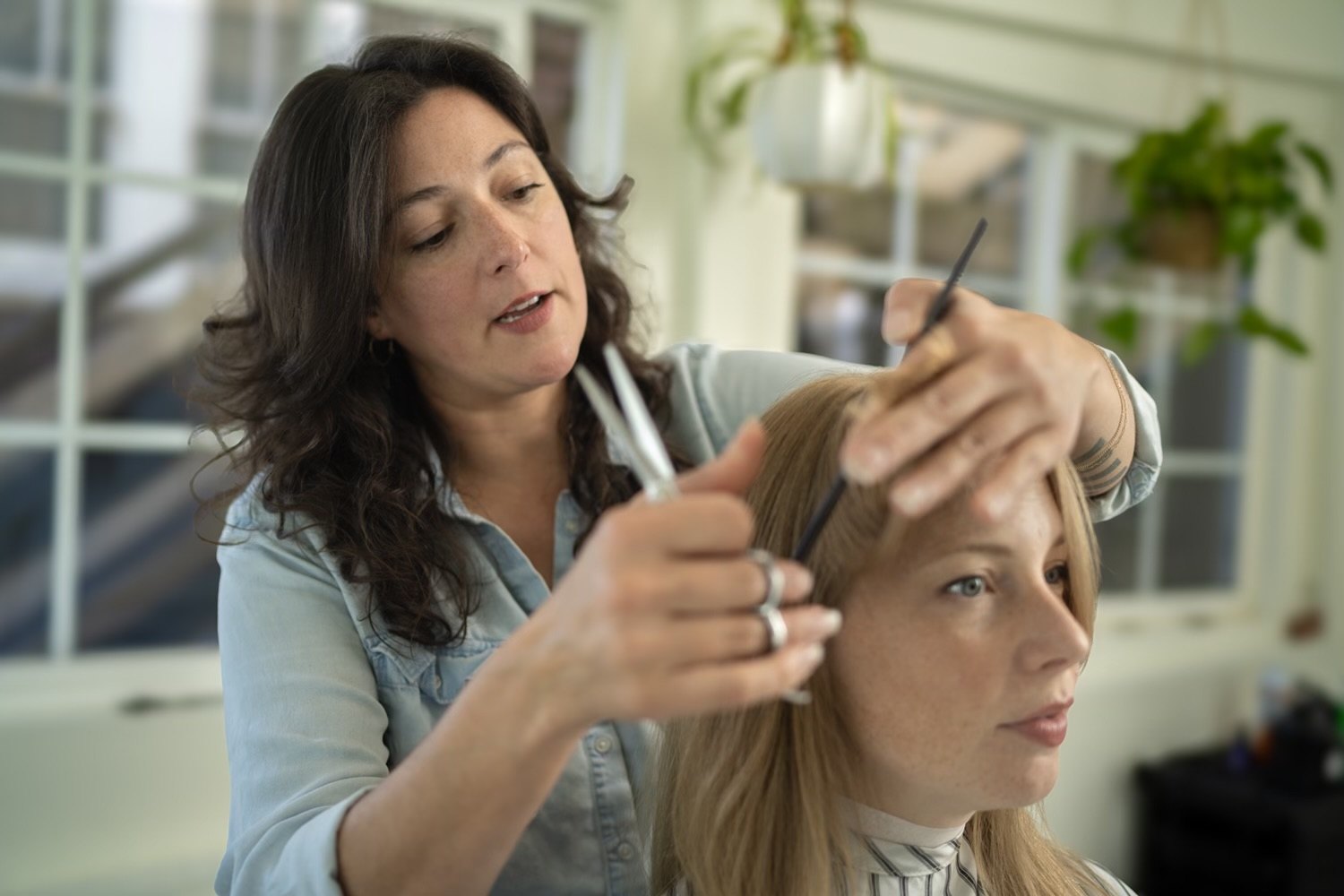 Natasha shaping a blonde client's hair with shears and a comb, talking her through it
